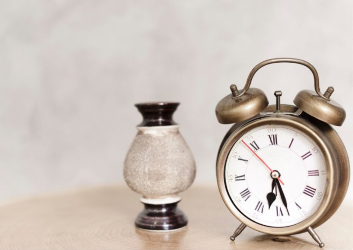 Gros plan sur une Horloge et Réveil vintage en laiton à côté d'un vase décoratif, sur une table en bois clair.